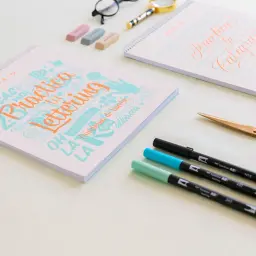 Cuadernos de "Practica Lettering" y "Practica tu Caligrafía" con rotuladores y tijeras doradas.
