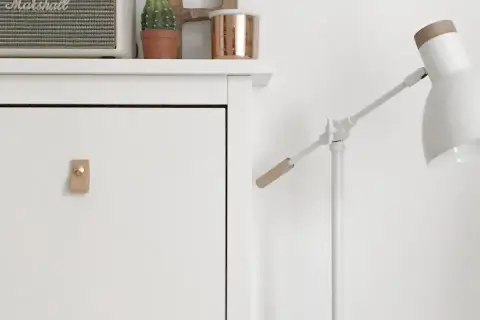 Un mueble blanco con un tirador de cuero y un altavoz Marshall. Una lámpara blanca con detalles de madera ilumina la escena.